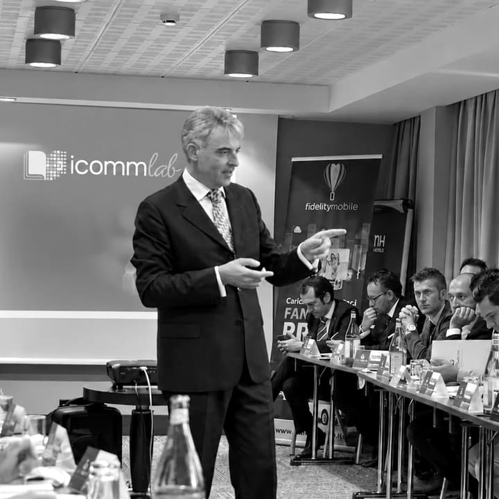 Giacomo Lenoci parla in piedi in una sala riunioni e indica le persone sedute al tavolo; alle sue spalle, lo schermo con il marchio di iComm Lab.